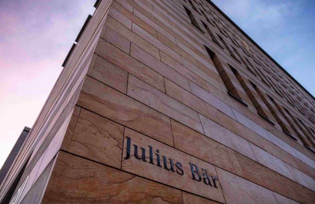 Edifício moderno de pedra com 'Julius Bär' na fachada. Vista de baixo para cima, céu colorido.