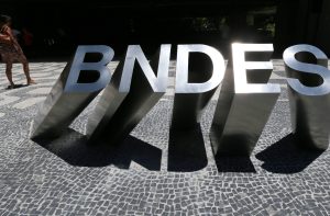 Letras metálicas "BNDES" em calçada, com sombras fortes e uma pessoa caminhando.