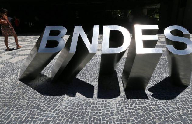 Letras metálicas "BNDES" em calçada, com sombras fortes e uma pessoa caminhando.