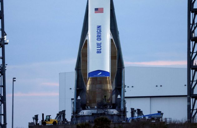 Foguete Blue Origin branco com bandeira dos EUA na plataforma de lançamento, ladeado por torre de serviço.