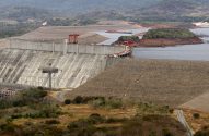 Enorme barragem de concreto com guindastes. Lago e montanhas ao fundo. Estrada sinuosa no terreno.