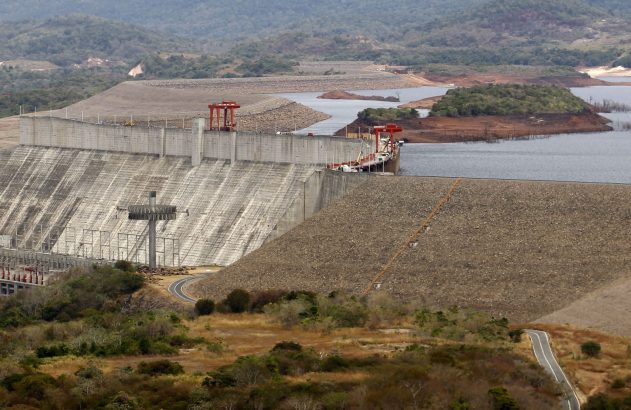 Enorme barragem de concreto com guindastes. Lago e montanhas ao fundo. Estrada sinuosa no terreno.