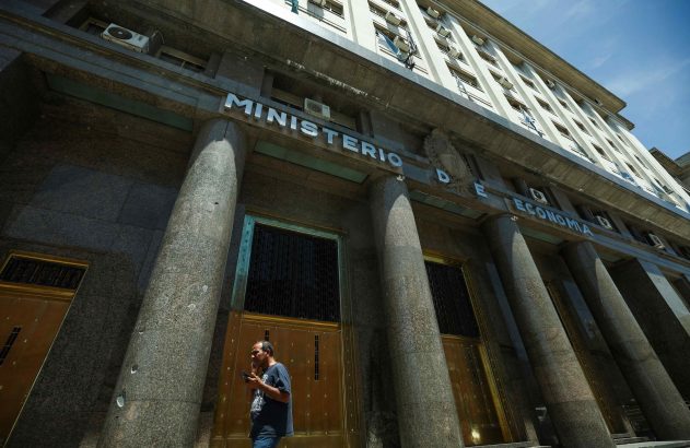 Homem olhando o celular em frente à imponente fachada do Ministério da Economia, com grandes colunas de granito e portas douradas.