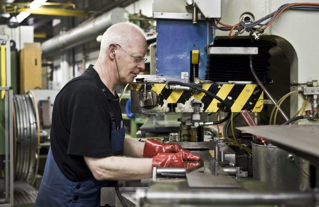 Operário com luvas vermelhas e fones trabalhando em prensa industrial, inserindo peça de metal.