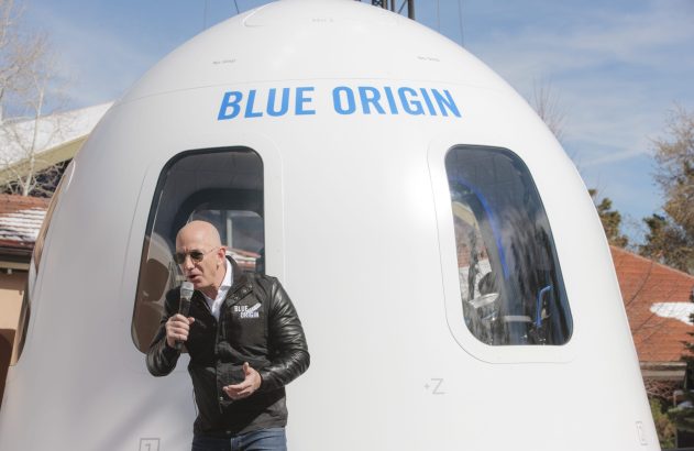 Homem careca em jaqueta Blue Origin fala ao microfone. Em frente, a cápsula branca 'BLUE ORIGIN' da empresa espacial.