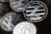 Várias moedas físicas de Litecoin, prateadas com logo 'L', sobre superfície escura e reflexiva.