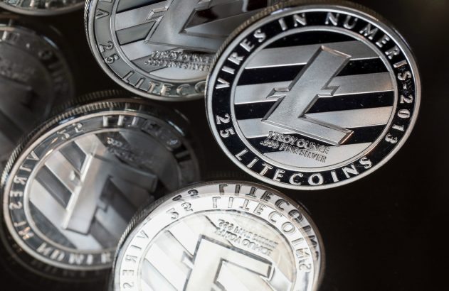 Várias moedas físicas de Litecoin, prateadas com logo 'L', sobre superfície escura e reflexiva.