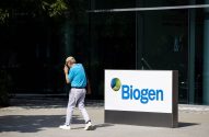Homem de boné com objeto na mão caminha em frente à placa da Biogen, com prédio envidraçado ao fundo.