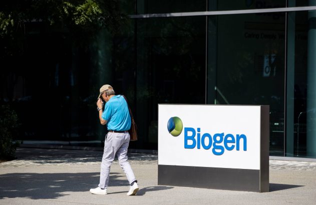 Homem de boné com objeto na mão caminha em frente à placa da Biogen, com prédio envidraçado ao fundo.