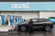 Pessoa carregando carro elétrico preto em estação Revel. Vários carregadores e um carro azul ao fundo.