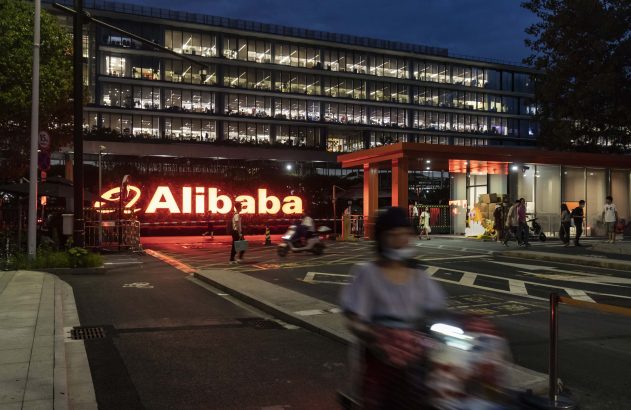 Sede da Alibaba iluminada à noite, com logo brilhante e pessoas na entrada.