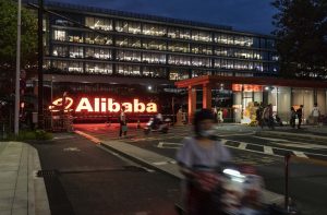 Sede da Alibaba iluminada à noite, com logo brilhante e pessoas na entrada.