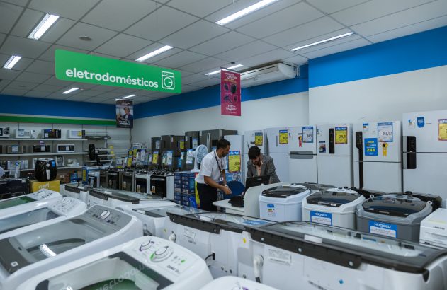 Dois homens interagem em balcão de loja de eletrodomésticos da Magalu, exibindo geladeiras e máquinas de lavar.
