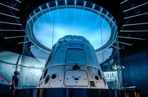 Cápsula espacial SpaceX em exibição, com iluminação azul e painéis de vidro. Um homem a observa.
