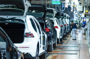 Linha de montagem de carros em fábrica, com veículos brancos e pretos, porta-malas abertos e trabalhadores.