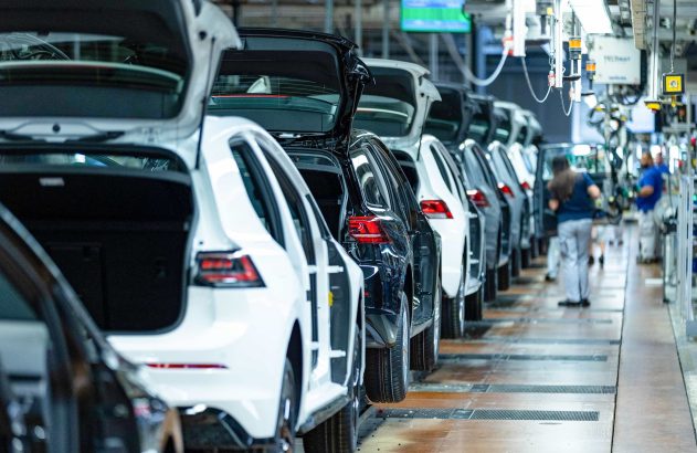 Linha de montagem de carros em fábrica, com veículos brancos e pretos, porta-malas abertos e trabalhadores.