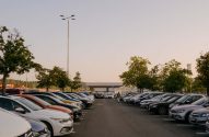 Estacionamento diurno com muitas fileiras de carros estacionados e uma pessoa caminhando em direção ao fundo.
