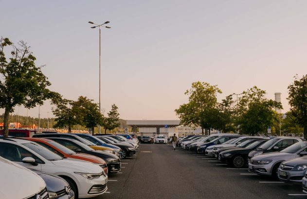 Estacionamento diurno com muitas fileiras de carros estacionados e uma pessoa caminhando em direção ao fundo.
