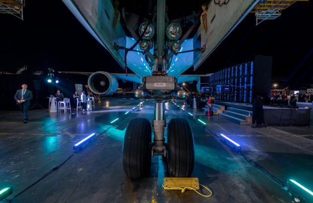Trem de pouso de avião iluminado por luzes azuis durante evento noturno, com pessoas em ambiente de hangar.