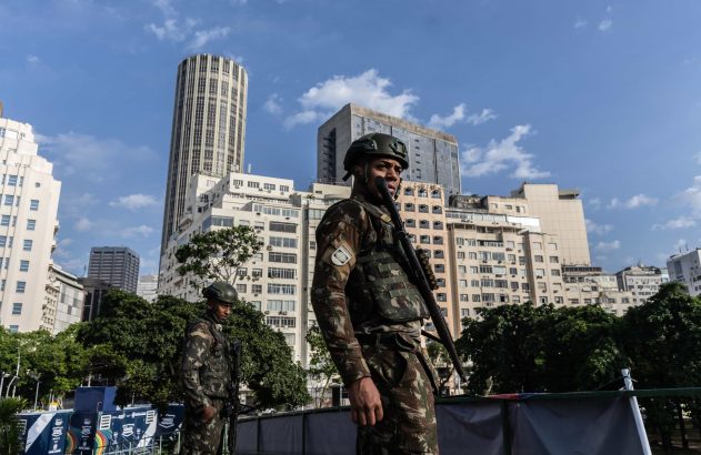 Dois soldados fardados em patrulha urbana. Um com escopeta. Ao fundo, prédios e banner G20.