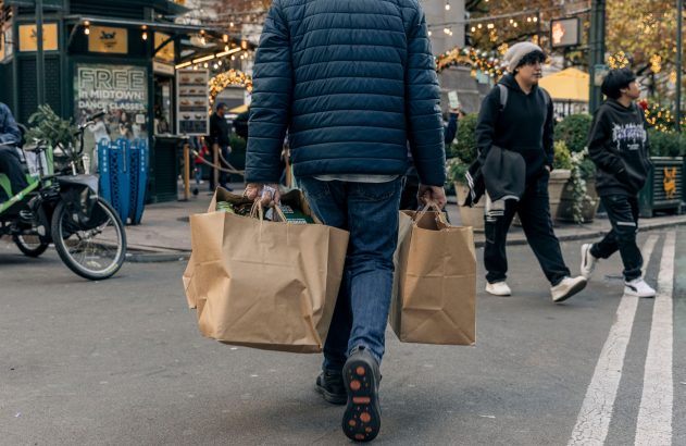 Pessoa de costas carregando sacolas de compras em rua urbana, com luzes e outras pessoas ao fundo.