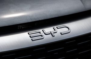 Logo metálico e brilhante da BYD na grade frontal de um carro cinza.