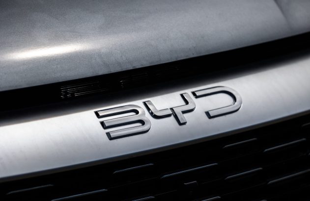 Logo metálico e brilhante da BYD na grade frontal de um carro cinza.