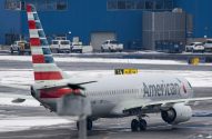 Avião da American Airlines taxiando em pista coberta de neve e gelo. Edifício azul e veículos no fundo.