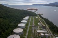 Terminal industrial com grandes tanques de armazenamento e navios petroleiros atracados em píer, rodeado por floresta e mar.