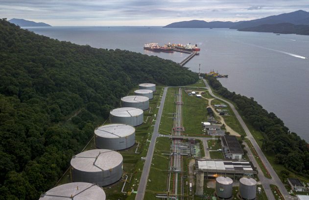 Terminal industrial com grandes tanques de armazenamento e navios petroleiros atracados em píer, rodeado por floresta e mar.