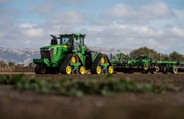 Trator John Deere verde de esteiras puxando implemento agrícola em campo arado sob céu nublado.
