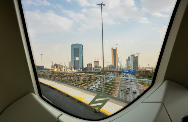 Vista de metrô elevado: cidade com arranha-céus, guindastes, rodovia com carros sob céu nublado.