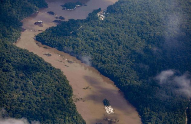 Vista aérea de rio barrento em floresta com fumaça, estruturas de mineração no rio e construções na margem.