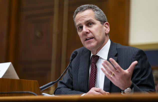 Um homem de cabelo grisalho, de terno e gravata, fala e gesticula em uma bancada com microfone.