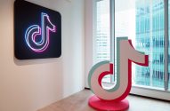 Dois logos do TikTok: um neon na parede e outro 3D no chão de um escritório moderno, com janela e prédios externos.