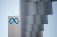 Logotipo azul do infinito da Meta em letreiro branco, com céu claro e estrutura desfocada ao lado.