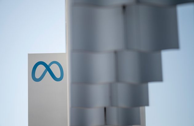 Logotipo azul do infinito da Meta em letreiro branco, com céu claro e estrutura desfocada ao lado.