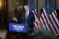 Donald Trump discursa em pódio "TRUMP VANCE" entre bandeiras dos EUA.
