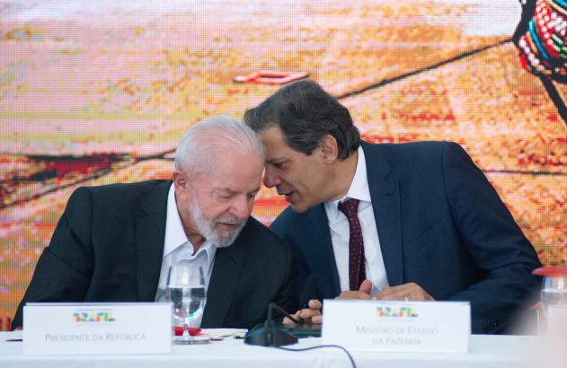 Presidente Lula e Ministro Haddad em mesa; Haddad sussurra para Lula. Tela colorida ao fundo.