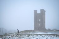 Pessoa e cachorro branco passeando na neve, com torre em nevoeiro ao fundo.