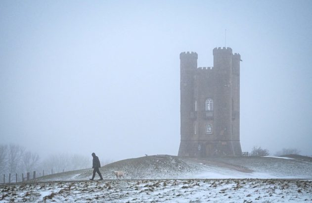 Pessoa e cachorro branco passeando na neve, com torre em nevoeiro ao fundo.