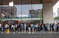 Longa fila de pessoas espera para entrar em uma Apple Store com fachada de vidro, em dia úmido.