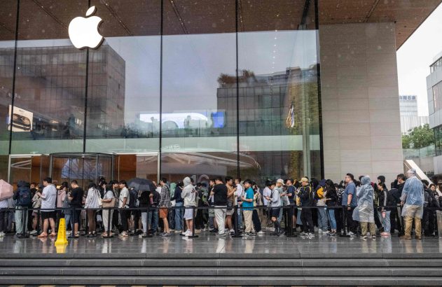 Longa fila de pessoas espera para entrar em uma Apple Store com fachada de vidro, em dia úmido.