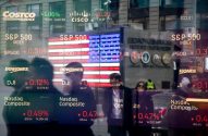 Reflexos de índices de mercado (S&P 500, Dow Jones, Nasdaq) em vitrine, com pessoas e bandeira dos EUA.