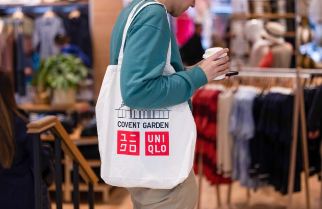 Pessoa em loja, segurando café e celular, com sacola branca Uniqlo Covent Garden.