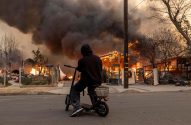 Pessoa em patinete elétrico, de costas, observa casas em chamas com fumaça escura ao fundo.