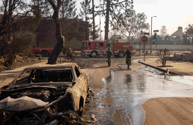 Consequências de incêndio: carro queimado, caminhão de bombeiros, 2 ciclistas em rua molhada e destruição.
