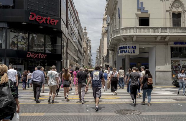 Multidão de pedestres atravessando faixa em rua comercial. Lojas Dexter e Librerias Levalle nas laterais.