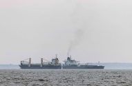 Duas embarcações: um cargueiro com peças e um petroleiro, que emite fumaça, cruzam-se no mar sob céu cinzento.