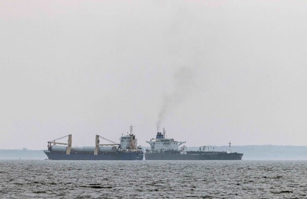 Duas embarcações: um cargueiro com peças e um petroleiro, que emite fumaça, cruzam-se no mar sob céu cinzento.